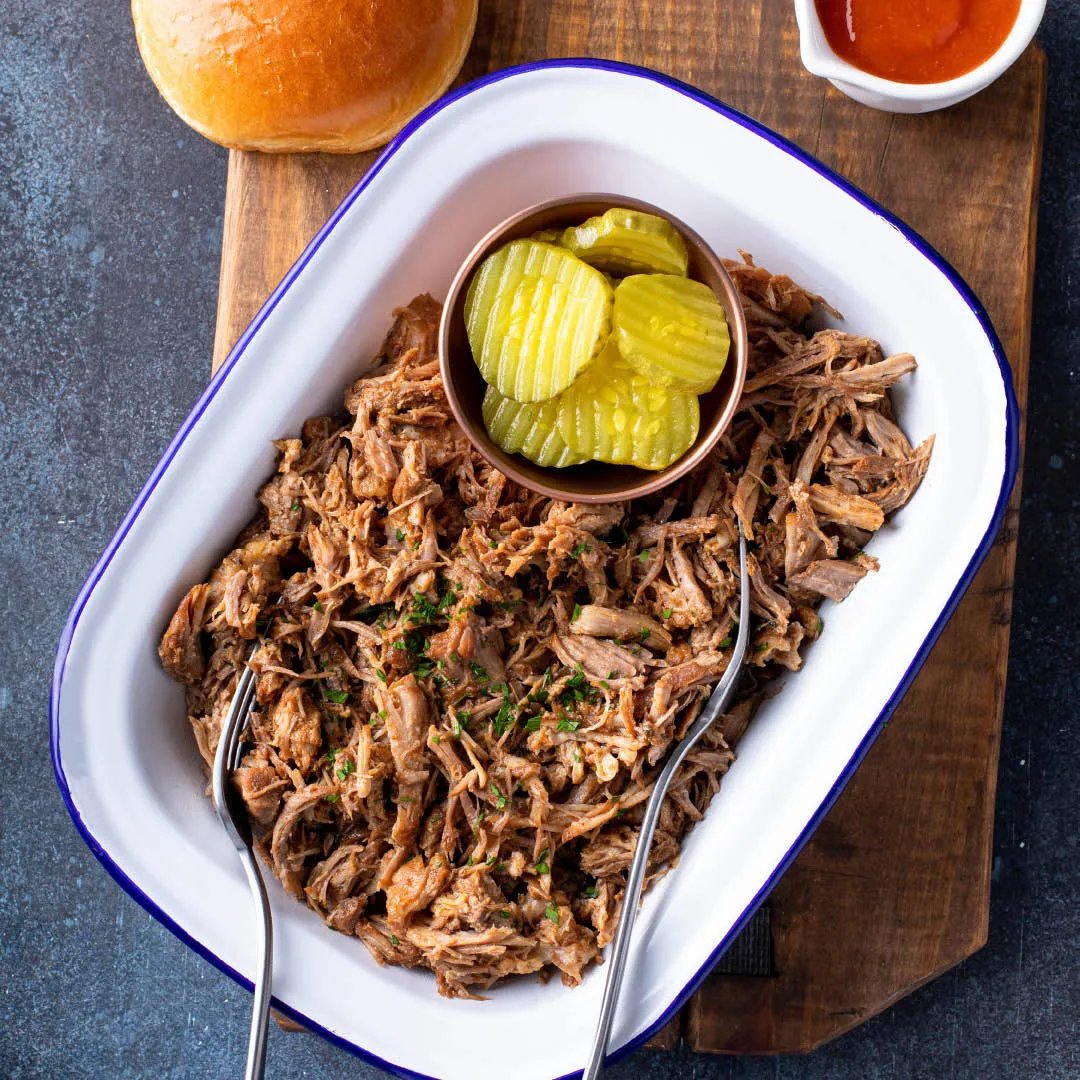 A white dish filled with shredded barbecue pulled pork, garnished with herbs, with a small bowl of sliced pickles on top. Two serving forks are in the dish. A bun and sauce are on a wooden board nearby.
